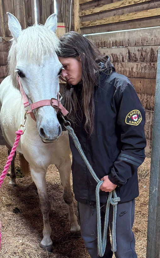 Person in black jacket embraces a white horse with a pink halter in a wooden stable, conveying a calm, affectionate mood.